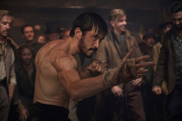 A shirtless, muscular man with dark hair and a beard holds a fighting pose with bandaged hands.