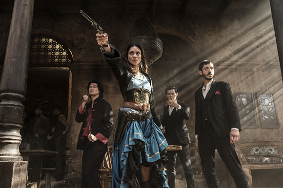 A woman in a corset and blue skirt holds a revolver up while four people stand behind her in a rustic building.