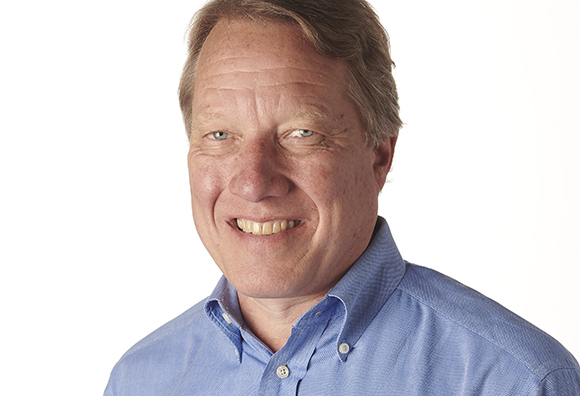 A smiling middle-aged man with light hair wearing a blue button-down shirt against a white background.
