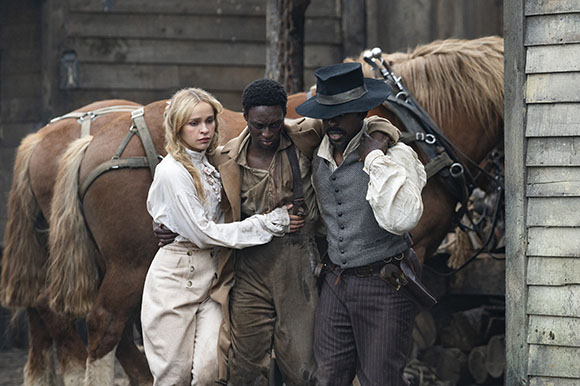 A blonde woman in white and a Black man in a tan shirt stand beside another Black man in a grey vest and hat.