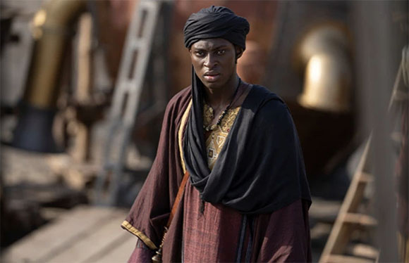 A person wearing a black headwrap and dark maroon and black robes standing outdoors.