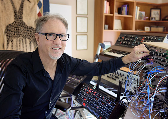 A man in a black shirt adjusts controls on a complex modular synthesizer with many patch cables.