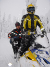 A person in a yellow and black snowsuit sits on a snowmobile in a snowy forest, with another person behind them.