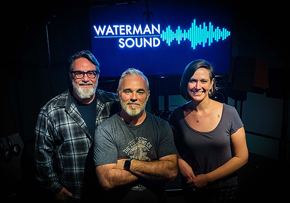 Three people smiling and posing in a dark studio in front of a blue Waterman Sound logo.