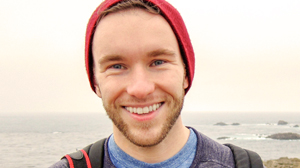 A smiling man with a beard wearing a red beanie outdoors near the ocean.