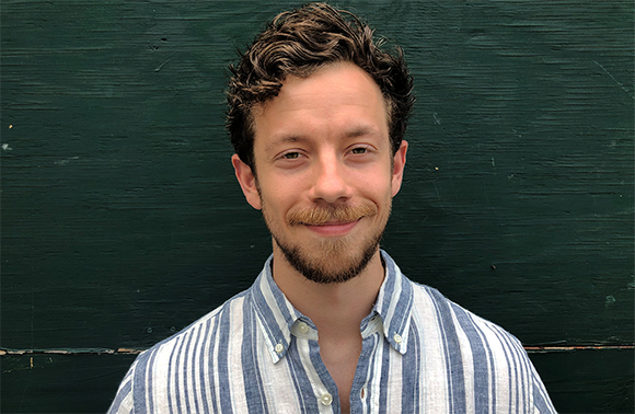 A man with curly brown hair and a beard smiling while wearing a blue and white striped shirt.