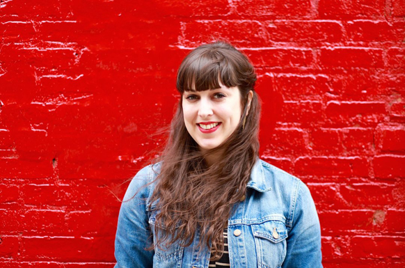 A woman with long brown hair and bangs wearing a denim jacket, smiling against a bright red brick wall.