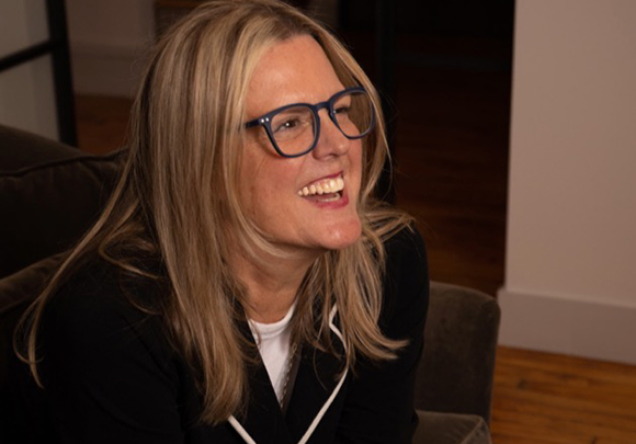A smiling blonde woman wearing blue-rimmed glasses and a black top.