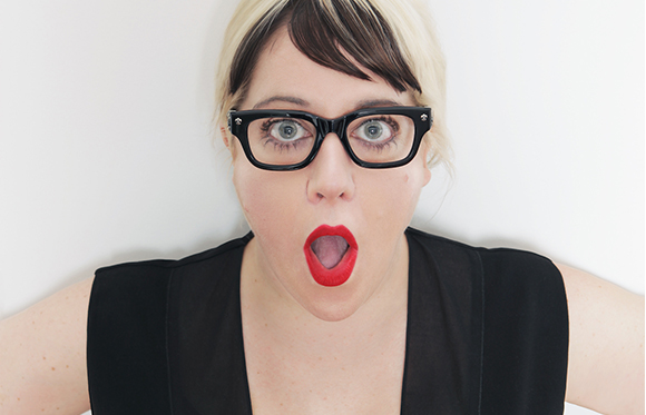 A woman with blonde hair and black glasses looks at the camera with a surprised expression and open mouth.