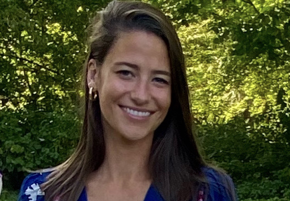 A smiling woman with long brown hair and gold hoop earrings stands before green trees.