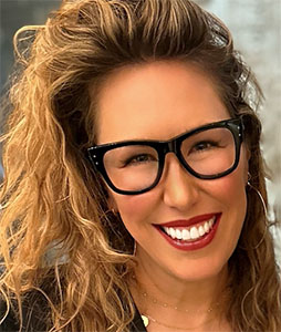 A smiling woman with curly blonde hair and black-rimmed glasses.
