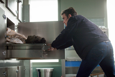 A man in a dark jacket leans over an open metal morgue drawer holding a deceased person.