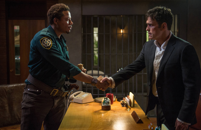 A man in a green uniform shakes hands with a man in a black suit across a wooden desk.