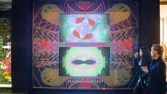 Two women stand in profile, observing a large, glowing digital art display with colorful abstract patterns.