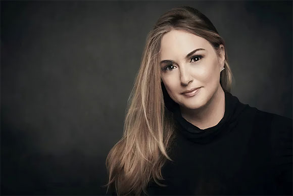 A woman with long blonde hair and a black turtleneck looks toward the camera against a dark background.