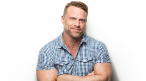 A man with short blond hair and a beard wearing a blue and white checkered short-sleeve shirt.