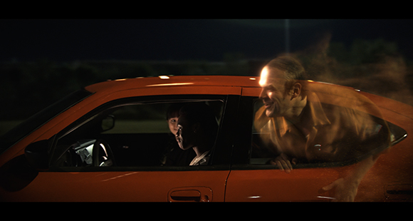 A translucent, glowing man leans against an orange car at night while two people sit inside.