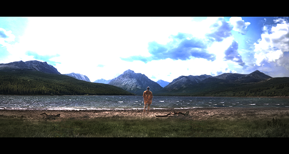 A person stands on a shoreline looking out over a large lake toward distant mountains under a cloudy sky.