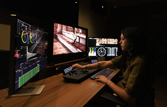 A person sits at a wooden desk using a keyboard and control panel with three large monitors displaying video data.
