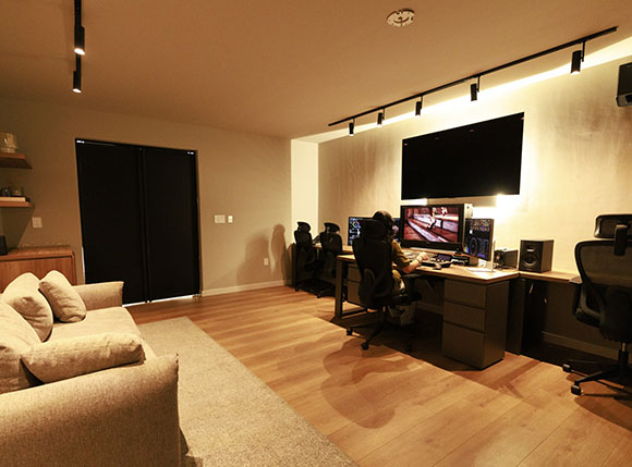 A gaming room featuring a long desk with multiple monitors, gaming chairs, and a beige sofa on a light wood floor.