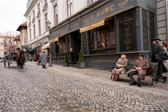 People in period clothing walk past a dark storefront labeled S. Kurc on a cobblestone street with a horse carriage.
