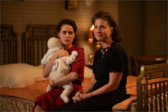 A woman in a red dress holds a baby while another woman in a black dress sits beside her on a bed.