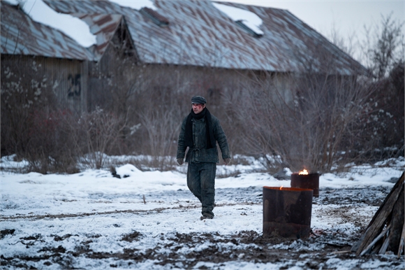 A man in a dark jacket and cap walks through a snowy field toward burning metal barrels.