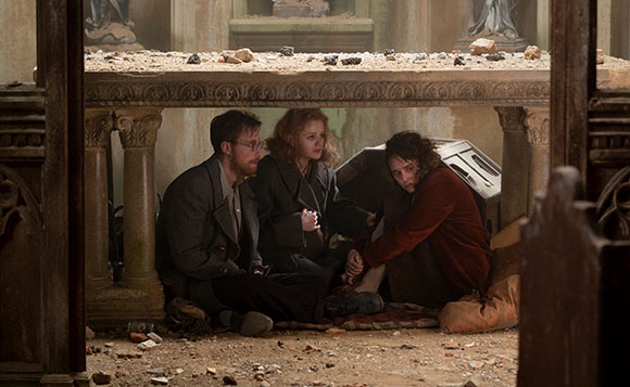 Three people sit huddled together on the floor of a damaged, stone building.