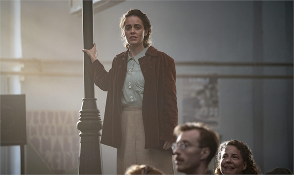 A woman in a brown jacket and light blue shirt stands by a lamppost with a worried expression.