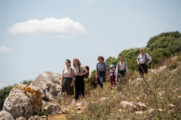 A group of people in vintage clothing hiking up a sunny, rocky hillside under a blue sky.