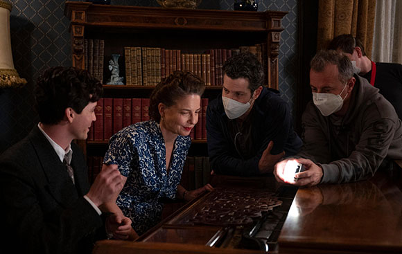 A man and woman in period clothing look at a piano while two crew members in masks inspect it with a light.