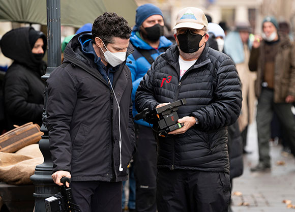 Two men in jackets and face masks stand outdoors on a city street near film equipment.