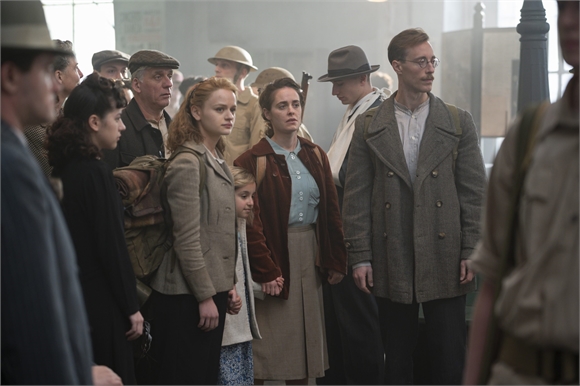 A group of people in mid-20th century clothing walking through a crowded street.