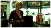 A blonde woman in a dark patterned shirt stands in a brightly lit cafe setting.