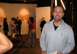 A man in a light blue button-down shirt stands in a studio with people and paintings in the background.