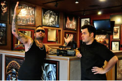 Two men, one in a black tank top and one in a black shirt, pose in a room with framed art.