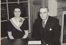 A man in a suit and glasses and a woman in a patterned dress sit side by side at a table.