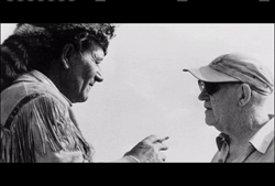 Black and white profile view of a man in fringed clothing and a man in a baseball cap facing one another.
