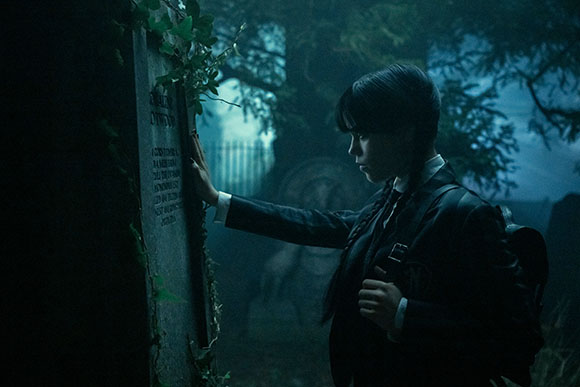 A young woman with dark braided hair touches a mossy tombstone in a dark, misty cemetery at night.