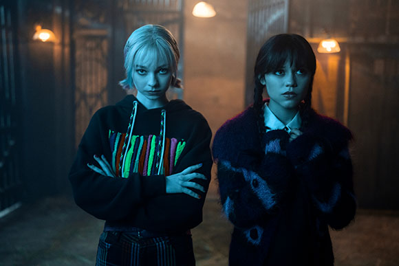 Two young women with serious expressions stand in a dark, moody room with warm overhead lighting.