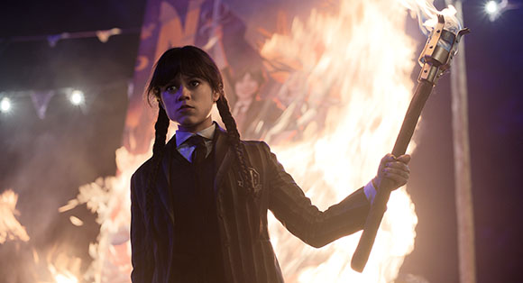 A young woman with dark braids holds a flaming torch in front of a large fire.
