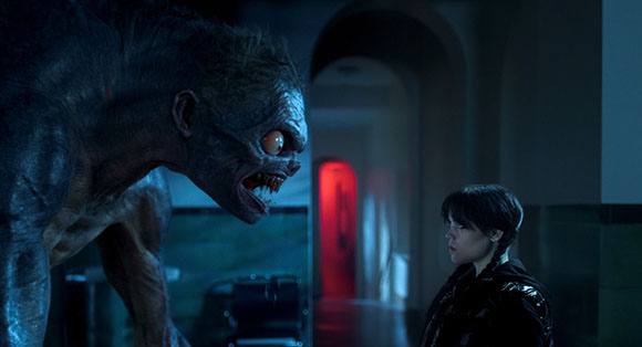 A large, dark, textured creature with large eyes leans toward a woman in a dimly lit hallway.
