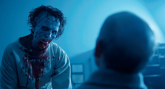 A pale, blood-streaked man with dark curly hair stares intensely at the back of a person's head in blue light.