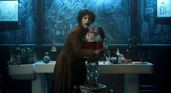 A man in a long brown coat holds a glass jar containing a brain in a dimly lit laboratory.