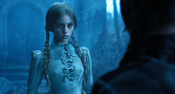 A young woman with blonde braids in a light-colored dress stares intensely in a blue-toned setting.