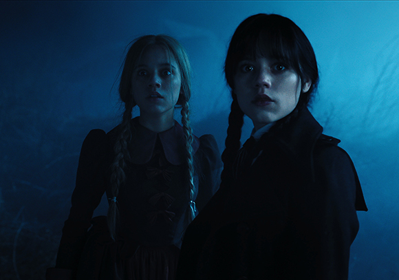 Two young girls with braided hair stand in a dark, blue-tinted setting, looking forward with serious expressions.