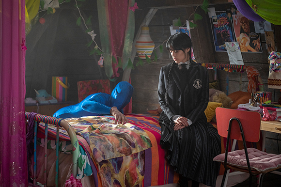 A girl in a black uniform sits on a bed next to a person in a blue bodysuit in a decorated room.