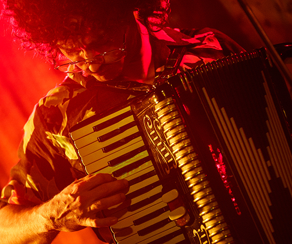 A person with curly hair plays an accordion under intense red stage lighting.