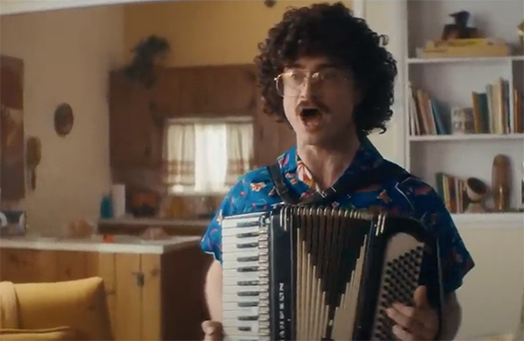 A man with curly hair and glasses playing an accordion while singing in a living room.