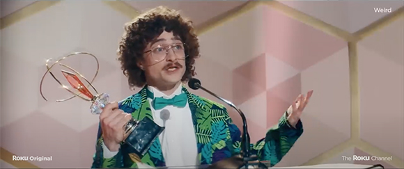 A man with curly hair, glasses, and a mustache wearing a tropical print blazer holds a gold trophy.